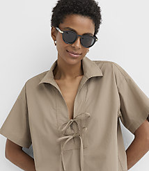 A woman with short, dark hair and sunglasses smiles against a plain white background, wearing a tan, collared shirt with a tie-front detail. carousel Product Image 2