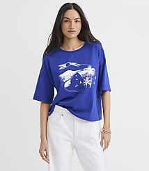A woman in a blue t-shirt with a white graphic of a tropical scene featuring palm trees and sailboats stands against a plain background. carousel Product Image 1