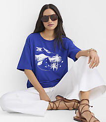 A woman in a blue graphic t-shirt and white pants sits in front of a plain white background. carousel Product Image 2