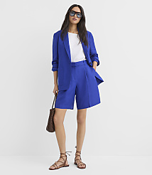 A woman in a blue blazer and shorts stands against a plain white background, holding a woven bag and wearing sandals. carousel Product Image 2