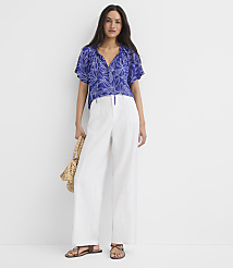 A woman in a blue floral top and white pants stands against a plain white background, holding a straw bag. carousel Product Image 2