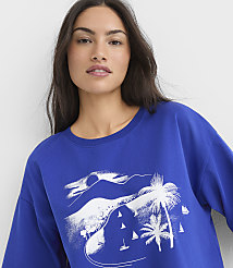 A woman in a blue sweatshirt with a white graphic of a tropical beach scene featuring palm trees and sailboats in the background. carousel Product Image 4