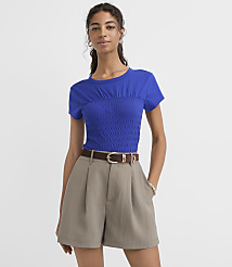 A woman in a blue smocked top and taupe shorts stands against a plain white background. carousel Product Image 1