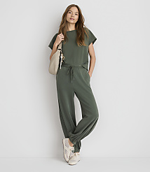 A woman in a green loungewear set and sneakers stands with a cream-colored bag slung over her shoulder against a plain white background. carousel Product Image 1