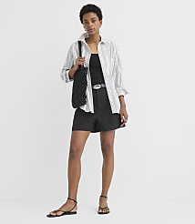 A woman in a striped shirt and black shorts stands against a plain white background, holding a black woven bag. carousel Product Image 3