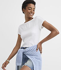 A smiling woman in a white smocked top and denim shorts is leaning against a light blue textured sweater against a plain white background. carousel Product Image 3