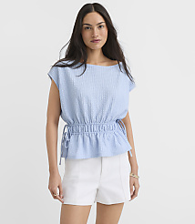 A woman in a light blue textured peplum top and white shorts stands against a plain white background. carousel Product Image 1