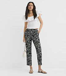 A woman in a white ruffled top and black leaf-patterned pants stands against a plain white background, holding a small white purse. carousel Product Image 1