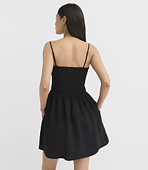 A woman in a black smocked-bodice mini dress with her hands in her pockets stands against a plain white background. carousel Product Image 3