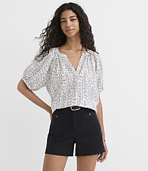 A woman with curly dark hair stands in the foreground wearing a patterned white blouse and black shorts, against a plain white background. carousel Product Image 1