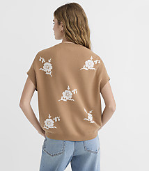 A woman in a tan, short-sleeved sweater with white floral embroidery stands against a plain white background. carousel Product Image 3