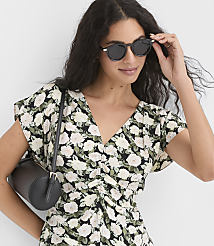 A woman in a floral dress and sunglasses adjusts her eyewear while a black cylindrical bag rests on her shoulder against a blurred background. carousel Product Image 1