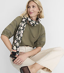 A blonde woman in a green top and cream pants, adorned with a floral scarf, sits against a plain white background. carousel Product Image 1