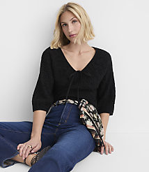 A woman in a black v-neck top and blue jeans sits against a plain white background. carousel Product Image 1