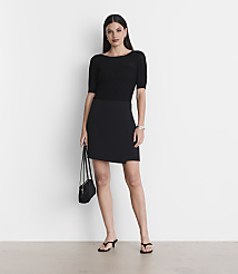 A woman in a black top and skirt stands against a plain white background, holding a black purse. carousel Product Image 1