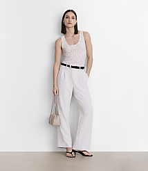 A woman in a white tank top and light-colored pants stands against a plain white wall, holding a small purse. carousel Product Image 1