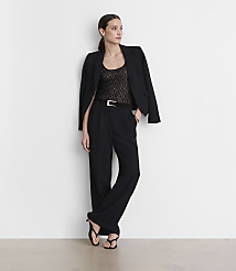 A woman in a black blazer, lace top, and wide-leg pants stands against a plain white wall.