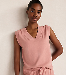 A young woman in a pink v-neck sweater vest stands against a textured, light-colored wall.