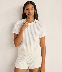 A woman in a cream-colored knit top and shorts stands against a textured, light-colored wall.