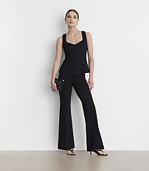 A woman in a black peplum top and flared pants stands against a plain white background, holding a black clutch.