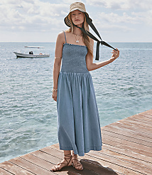 A woman in a blue dress and straw hat stands on a wooden pier with a boat in the calm ocean behind her. carousel Product Image 1