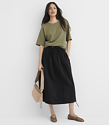 A woman in a green t-shirt and black skirt stands against a white background, holding a tan crossbody bag. carousel Product Image 2