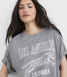 A woman in a grey t-shirt with a tiger graphic and "Los Angeles California" text stands against a plain white background. carousel Product Image 1