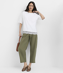 A woman in a white top with lace trim and olive green pants stands against a plain white background, holding a brown purse. carousel Product Image 2