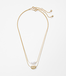 Two delicate gold necklaces, one with a white stone and the other with a gold pendant, lie on a plain white background. carousel Product Image 1