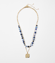 A delicate gold necklace with blue beads and a square pendant rests on a plain white background. carousel Product Image 1