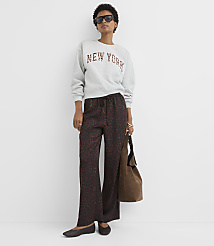 A woman in a "New York" sweatshirt and patterned pants stands against a plain white wall, holding a brown bag. carousel Product Image 3