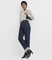 A woman in a striped sweater, dark jeans, and black boots stands against a plain white background, holding a black purse. carousel Product Image 2