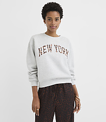 A woman in a gray sweatshirt with "NEW YORK" in leopard print stands against a plain gray background. carousel Product Image 2