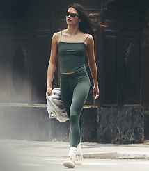 A woman in a green athletic outfit walks across a street with a blurred, dark building in the background. carousel Product Image 1
