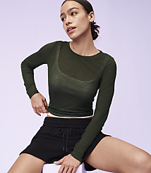 A woman in a sheer green long-sleeved shirt and black shorts sits against a soft purple background. carousel Product Image 4