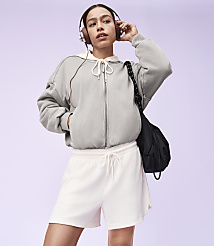 A young woman in a grey zip-up hoodie and white shorts stands with headphones on, a black bag slung over her shoulder, against a soft purple background. carousel Product Image 4
