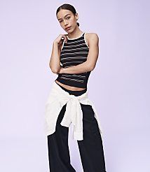 A woman in a striped tank top and black pants stands against a plain purple background, with a white sweater tied around her waist. carousel Product Image 4