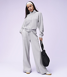 A young woman in a light gray tracksuit and white headband stands with a black bag in her right hand against a soft purple background. carousel Product Image 3