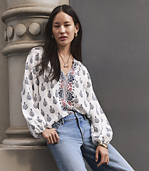A woman in a patterned blouse and jeans leans against a textured wall with a decorative column in the foreground. carousel Product Image 1