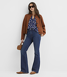 A woman in a brown cardigan, floral blouse, and flared jeans stands against a plain white background, holding a small brown purse. carousel Product Image 1