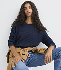 A woman with long, dark, curly hair wears a navy blue textured sweater and jeans, with a tan jacket draped over her shoulders, against a plain white background. carousel Product Image 1