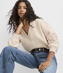 A woman in a beige v-neck sweater and blue jeans poses against a plain white background. carousel Product Image 1