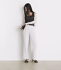 A woman in a black lace top, white pants, and a light cardigan stands against a plain white wall, holding a small black clutch. carousel Product Image 1