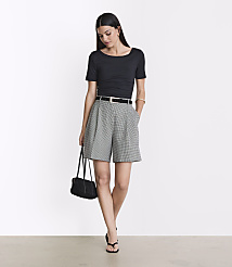 A woman in a black top and houndstooth shorts stands against a plain white background, holding a black purse. carousel Product Image 1