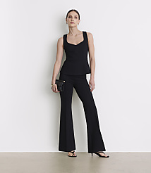 A woman in a black peplum top and flared pants stands against a plain white background, holding a black clutch.
