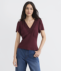 A woman in a burgundy v-neck top with lace trim and blue jeans stands against a plain white background. carousel Product Image 2