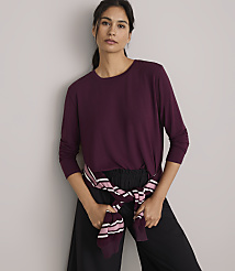 A woman in a burgundy long-sleeved shirt and black pants stands against a plain gray background, with a striped sweater tied around her waist. carousel Product Image 1