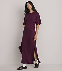 A woman in a plum-colored maxi dress with a side slit stands against a plain, light gray background, holding a dark bag. carousel Product Image 1