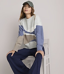 A woman in a patterned sweater and navy pants sits on a white chair against a plain white background. carousel Product Image 1