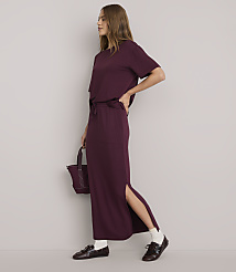 A woman in a burgundy t-shirt and matching maxi skirt with a side slit stands against a plain white background, holding a small burgundy bag.